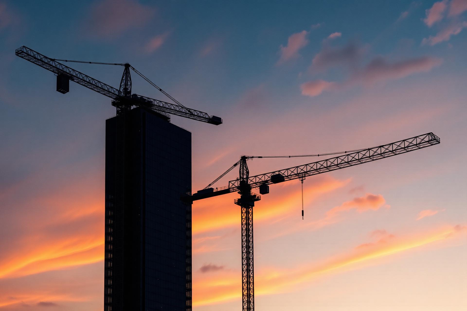 Cranes silhouetted against a dusk sky over a tower under construction
